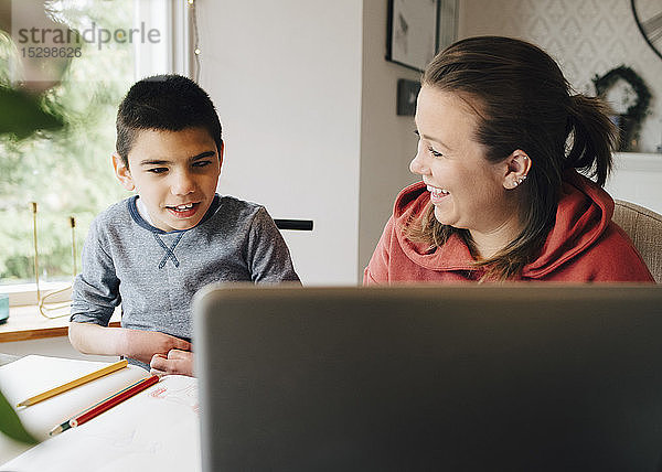 Lächelnde Mutter sieht autistischen Sohn an  der sich im Wohnzimmer ein Video über einen Laptop ansieht