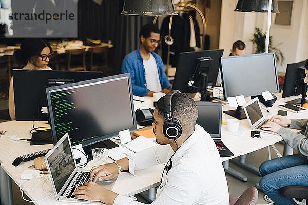 Hochwinkelansicht eines Geschäftsmannes mit Kopfhörer-Codierung über Laptop  während seine Kollegen im Hintergrund arbeiten