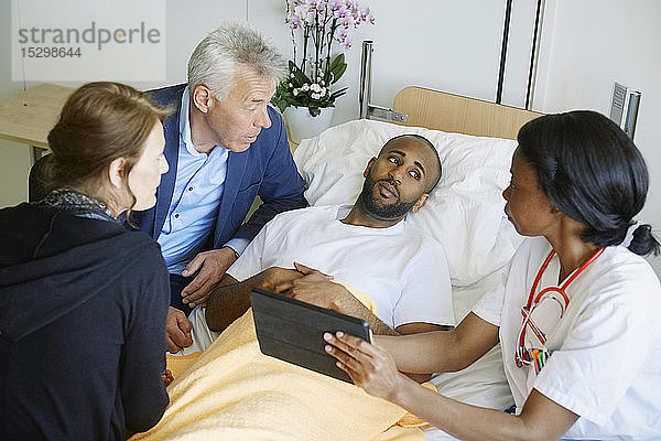 Hochwinkelansicht eines Arztes  der über ein digitales Tablett mit Patient und Familie auf der Krankenhausstation diskutiert