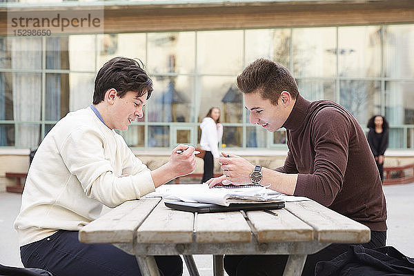 Männliche Freunde im Teenageralter lächeln beim Lernen am Tisch auf dem Schulhof
