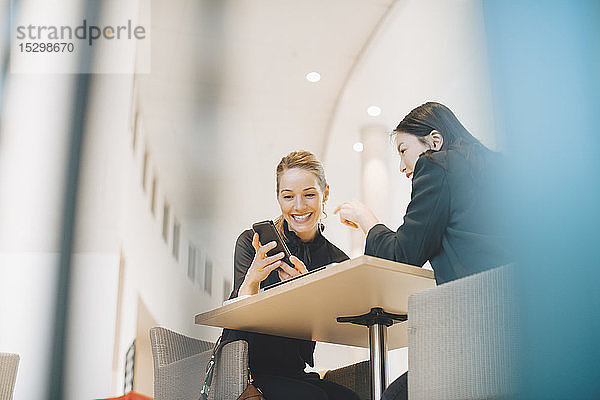 Niedriger Blickwinkel auf eine lächelnde Geschäftsfrau  die ihr Smartphone mit einer Tischkollegin im Büro teilt