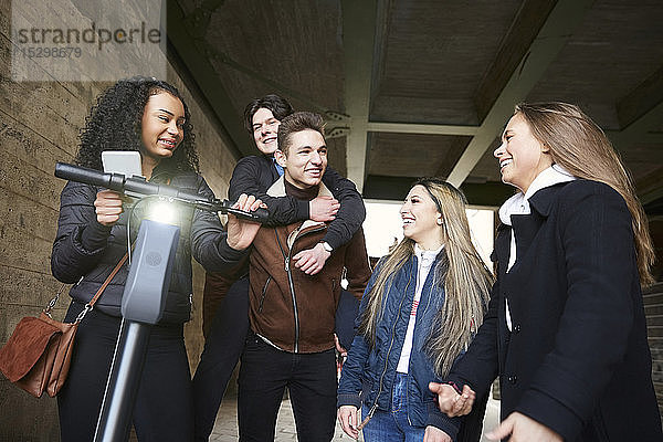 Lächelnde männliche und weibliche Teenager-Freunde mit Elektro-Schieberoller auf dem Fußweg unterhalb der Brücke