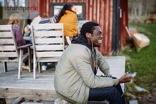 Fröhlicher junger Mann benutzt Smartphone  während sich Freunde bei Sonnenuntergang im Hintergrund unterhalten