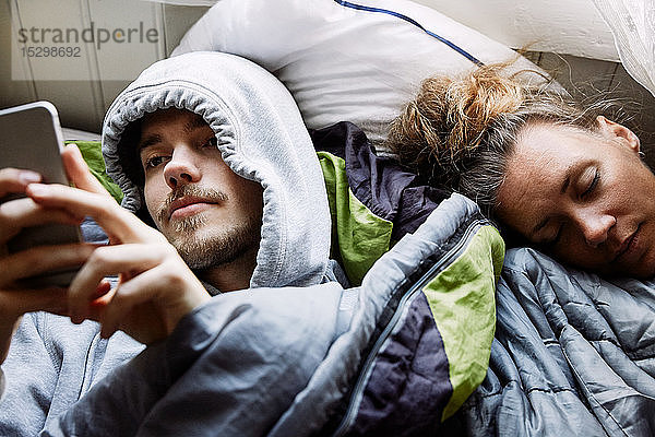 Schrägansicht eines jungen Mannes  der ein Smartphone benutzt  während er bei einem Freund zu Hause im Bett liegt
