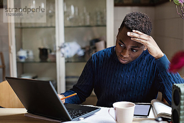 Besorgter Teenager schaut auf den Laptop  während er auf dem Tisch Hausaufgaben macht