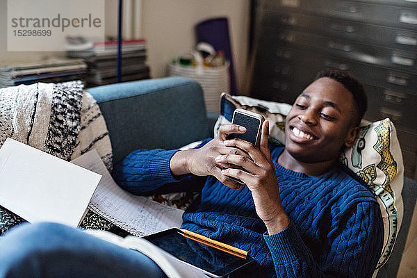 Lächelnder Teenager nutzt soziale Medien  während er bei den Hausaufgaben im Wohnzimmer auf dem Sofa liegt