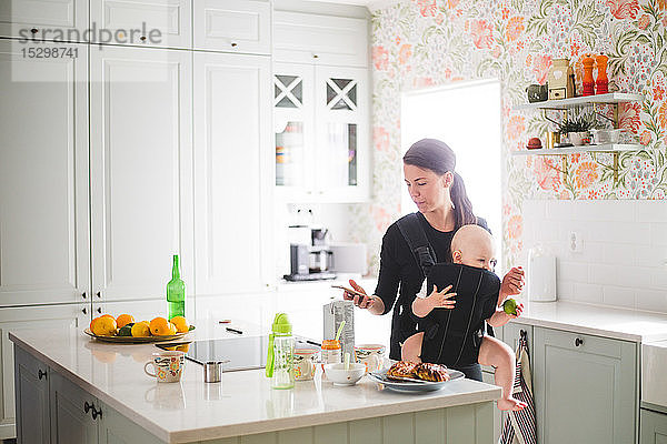 Mutter benutzt Mobiltelefon  während sie ihre Tochter in der Küche zu Hause trägt