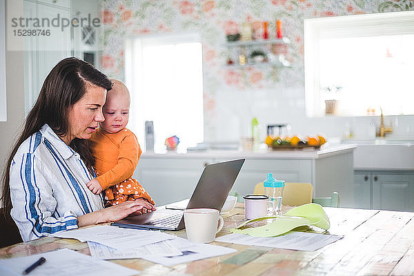 Unternehmerin  die eine liebende Tochter trägt  während sie ihren Laptop auf dem Esstisch im Heimbüro benutzt
