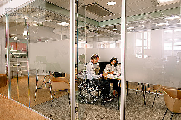 Geschäftsmann sitzt im Rollstuhl  während er mit einer Kollegin im Sitzungssaal am Arbeitsplatz mit Blick durch die Tür arbeitet