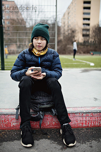 Junge in voller Länge mit Handy  der im Winter gegen den Fußballplatz schaut