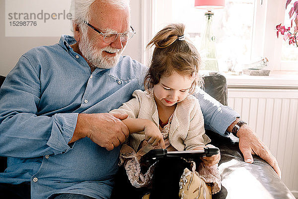 Mädchen benutzt digitales Tablet  während sie mit Großvater zu Hause auf dem Sofa sitzt