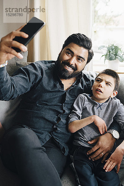 Lächelnder Vater  der mit seinem autistischen Sohn Selbsthilfe nimmt  während er auf dem Sofa im Wohnzimmer sitzt