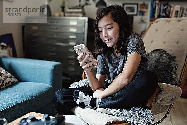 Lächelnder Junge benutzt Mobiltelefon  während er auf einem Stuhl im Wohnzimmer sitzt