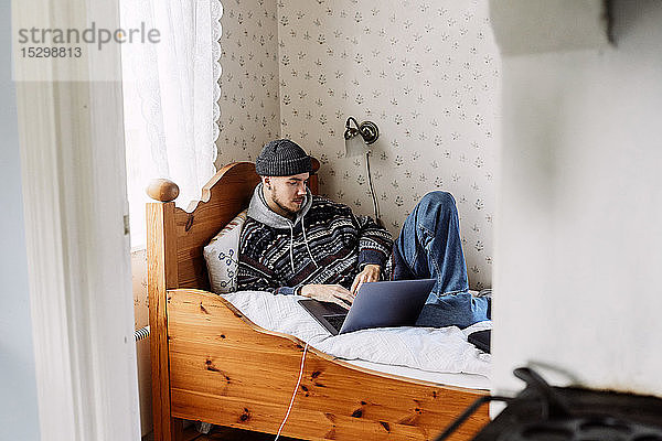 Junger Mann benutzt Laptop  während er sich auf dem Bett ausruht und durch die Tür zu Hause gesehen wird