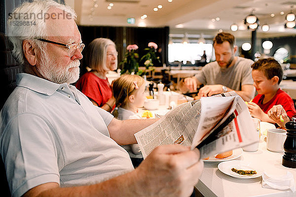 Älterer Mann liest Zeitung  während er mit seiner Familie beim Frühstück im Restaurant sitzt
