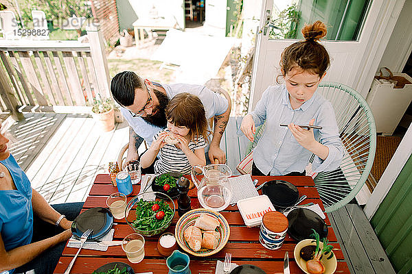 Schrägaufnahme eines Mädchens  das ein Mobiltelefon benutzt  während es mit seiner Familie zu Hause zu essen bekommt