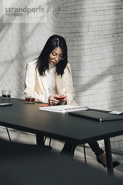 Mittlere erwachsene Geschäftsfrau benutzt Mobiltelefon am Tisch im Büro