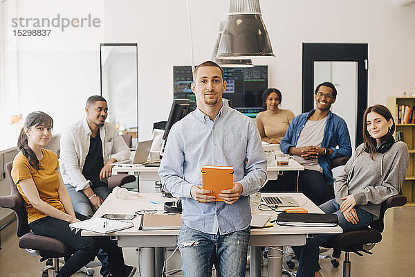 Porträt eines selbstbewussten Geschäftsmannes  der ein Buch in der Hand hält  während er mit Kollegen im Hintergrund im Kreativbüro sitzt
