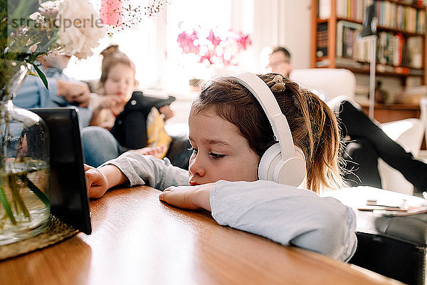 Mädchen hört Musik über Kopfhörer und benutzt ein digitales Tablet mit der Familie im Hintergrund