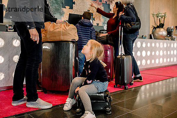 Mädchen sitzt auf einem Koffer und sieht den Vater an  während die Familie im Hintergrund steht