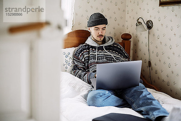 Junger Mann benutzt Laptop  während er entspannt auf dem Bett liegt und durch die Tür zu Hause gesehen wird