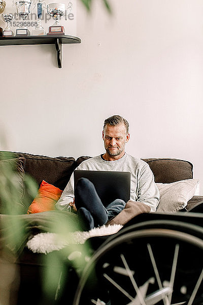 Behinderter reifer Mann benutzt Laptop  während er zu Hause auf dem Sofa sitzt