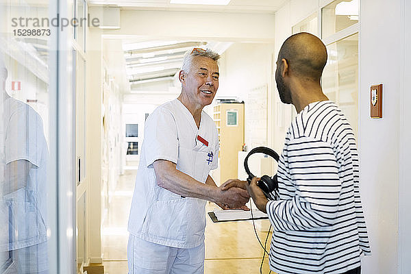 Leitender Arzt mit Klemmbrett begrüßt jungen Mann im Krankenhauskorridor