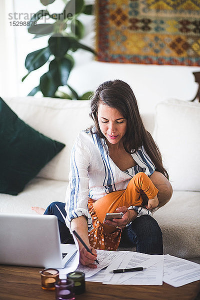 Freiberufliche Mitarbeiterin erledigt Papierkram  während sie ihre Tochter zu Hause auf dem Sofa stillt