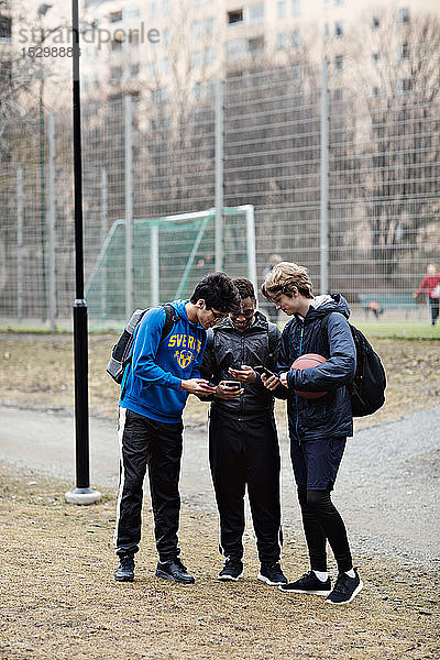 Social-Media-süchtige Freunde in voller Länge  die Sportkleidung tragen  während sie auf dem Spielfeld Mobiltelefone benutzen