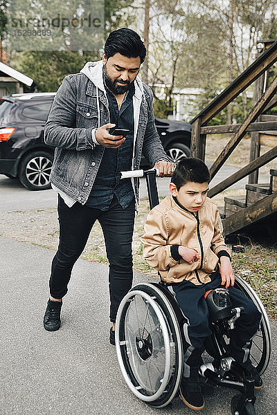 Vater benutzt Smartphone beim Schieben eines autistischen  im Rollstuhl sitzenden Sohnes