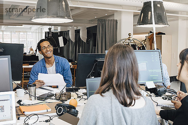 Fröhliche multiethnische IT-Fachleute unterhalten sich am Schreibtisch in einem kreativen Büro