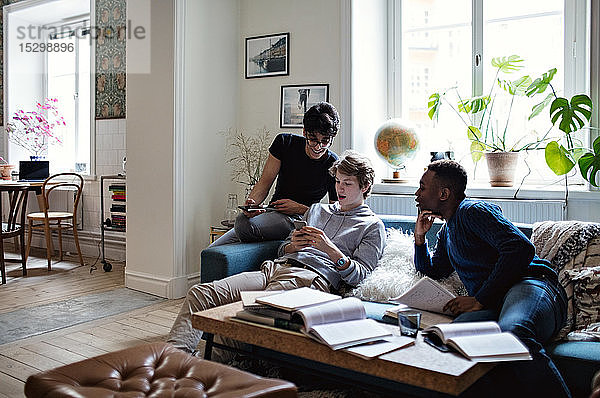 Männliche Freunde benutzen Mobiltelefone  während sie bei den Hausaufgaben im Wohnzimmer auf dem Sofa sitzen