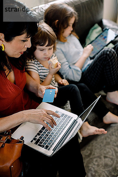 Schrägansicht einer Frau beim Online-Shopping mit einem Laptop  während sie mit ihren Töchtern zu Hause sitzt