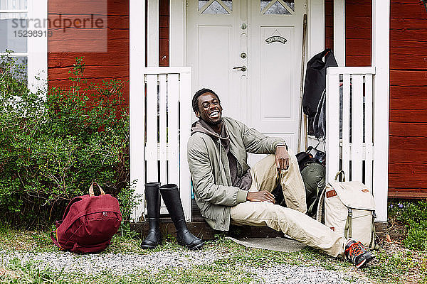 Porträt eines glücklichen jungen Mannes mit Rucksäcken und Stiefeln vor einem Ferienhaus sitzend
