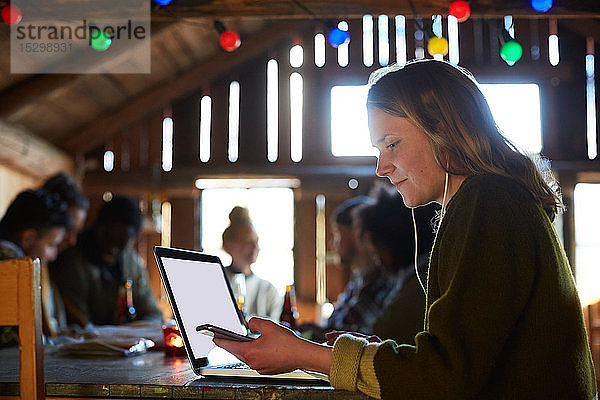 Junge Frau benutzt Laptop auf dem Tisch  während sich Freunde zu Hause im Hintergrund unterhalten