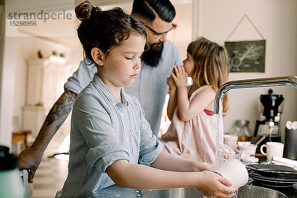 Mädchen wäscht Becher am Waschbecken  während der Vater in der Küche mit der Tochter spricht
