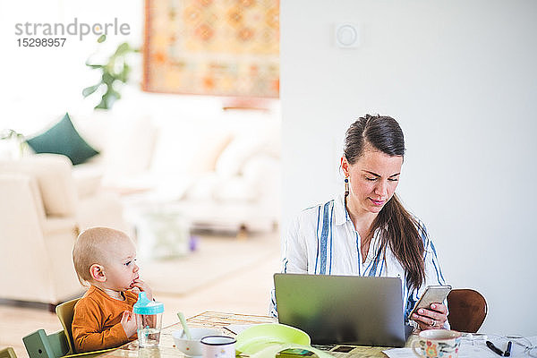 Kleinkind schaut der fleißigen berufstätigen Mutter zu  während es im Heimbüro am Tisch sitzt