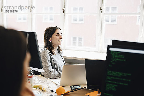 Lächelnde Computerprogrammiererin schaut weg  während sie im Büro am Schreibtisch sitzt