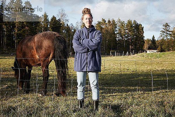 Porträt einer selbstbewussten  mittelgroßen  erwachsenen Frau mit verschränkten Armen  die zu Pferd auf einem Grasfeld gegen den Himmel steht