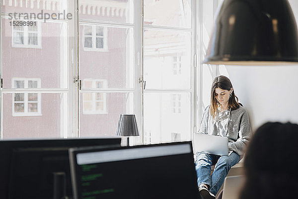 Fokussierte junge Programmiererin  die einen Laptop benutzt  während sie im Kreativbüro am Fenster sitzt