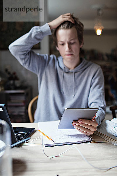 Müder Teenager mit Hand in den Haaren  der ein digitales Tablett benutzt  während er zu Hause Hausaufgaben auf dem Tisch macht