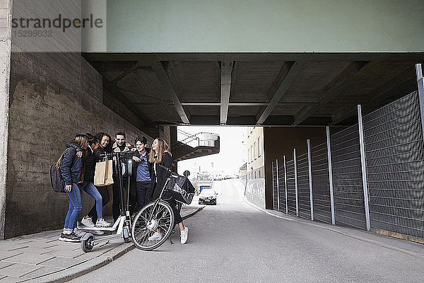 Männliche und weibliche Freunde im Teenageralter mit Roller und Fahrrad auf dem Bürgersteig unter der Brücke