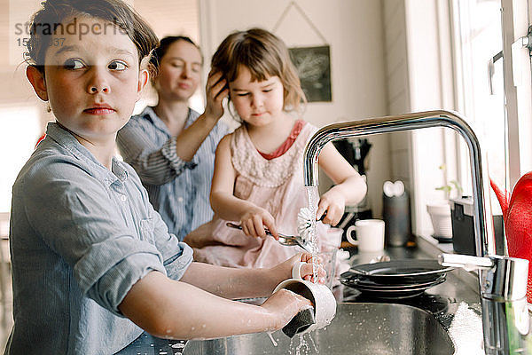 Mädchen wäscht Becher an der Küchenspüle mit Schwester und Mutter im Hintergrund zu Hause