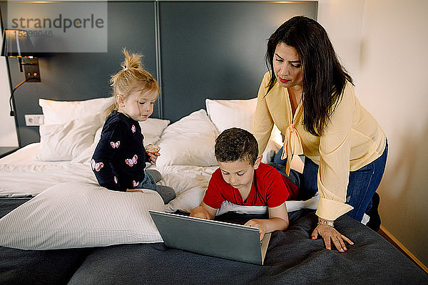 Mutter und Mädchen mit einem Jungen  der einen Laptop am Bett im Hotelzimmer benutzt