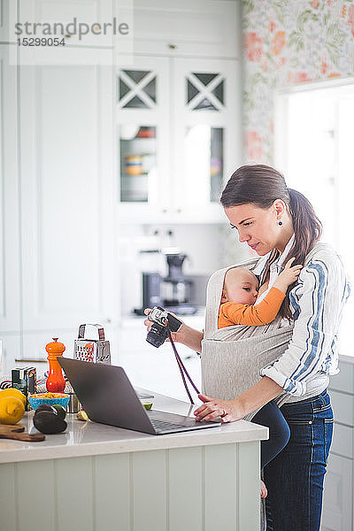 Seitenansicht einer Lebensmittel-Bloggerin mit Kamera  die einen Laptop benutzt  während sie ein kleines Mädchen in der Küche trägt