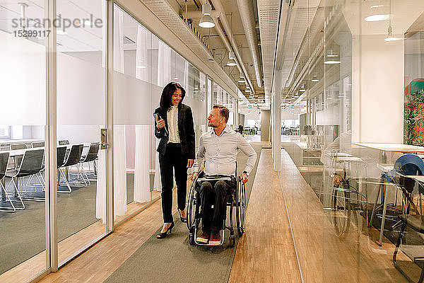 Lächelnde Geschäftsfrau betrachtet behinderte Kollegin  die am Arbeitsplatz im Rollstuhl auf dem Flur sitzt