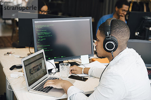 Seitenansicht eines männlichen Programmierers mit Kopfhörer-Codierung über Laptop auf dem Schreibtisch  während er im Büro sitzt
