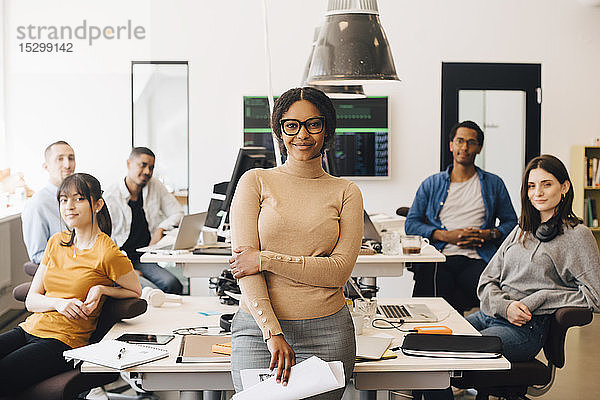 Porträt einer lächelnden Geschäftsfrau  die mit selbstbewussten Kollegen am Schreibtisch in einem kreativen Büro steht