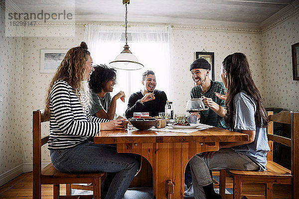 Glückliche Freunde unterhalten sich  während sie zu Hause Essen und Trinken auf dem Tisch genießen