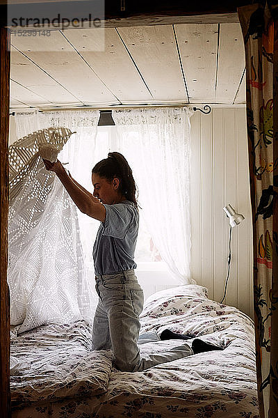 Seitenansicht einer jungen Frau  die ihre Decke zusammenlegt  während sie im Ferienhaus auf dem Bett kniet